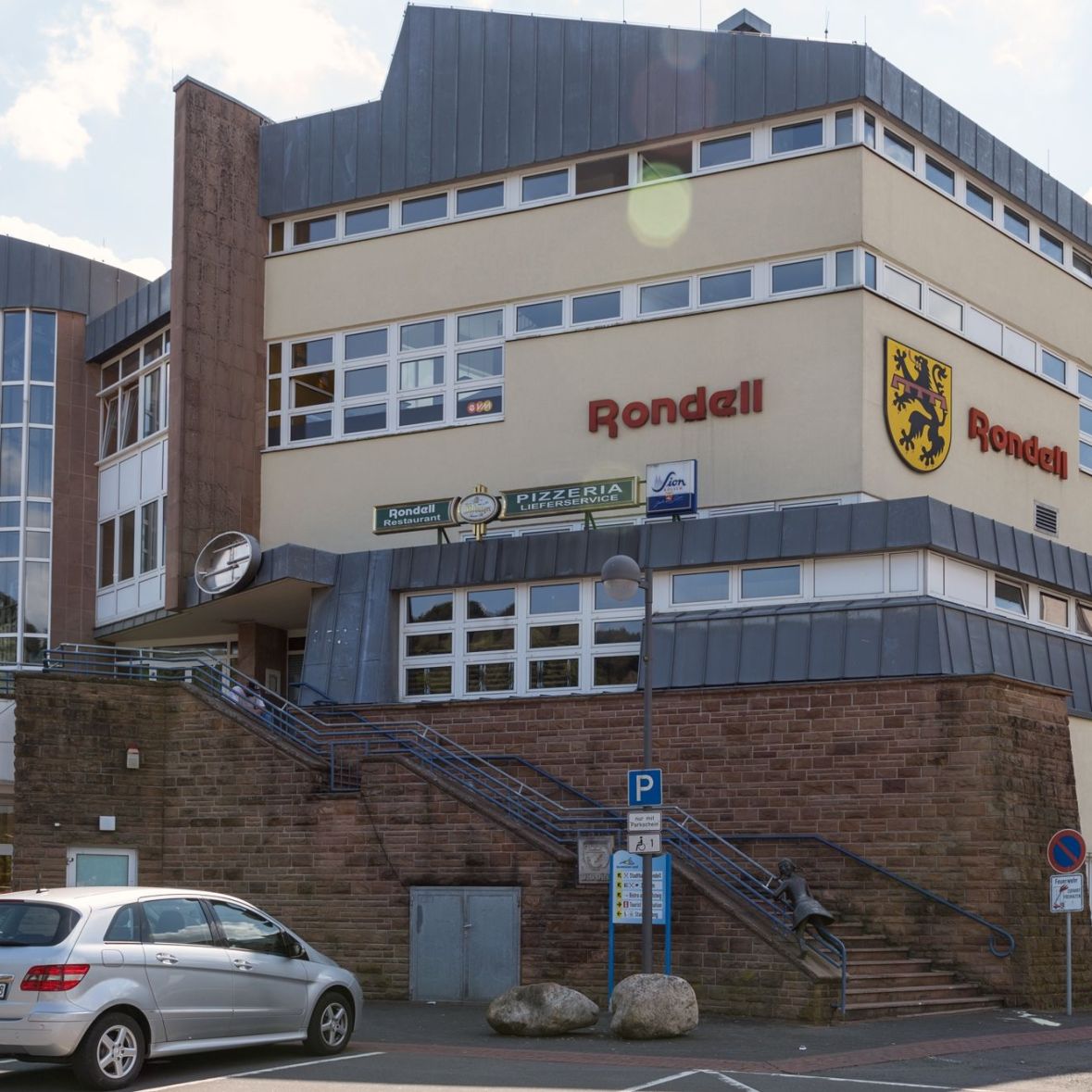 evm-Kundenzentrum Gerolstein, Ansicht von außen. Ein beige-gelbes Gebäude mit dem Schriftzug "Rondell" und einem Löwenwappen, Treppen führen hinauf, umgeben von Straßenverkehrsschildern und einem "NORMA"-Laden im Vordergrund.