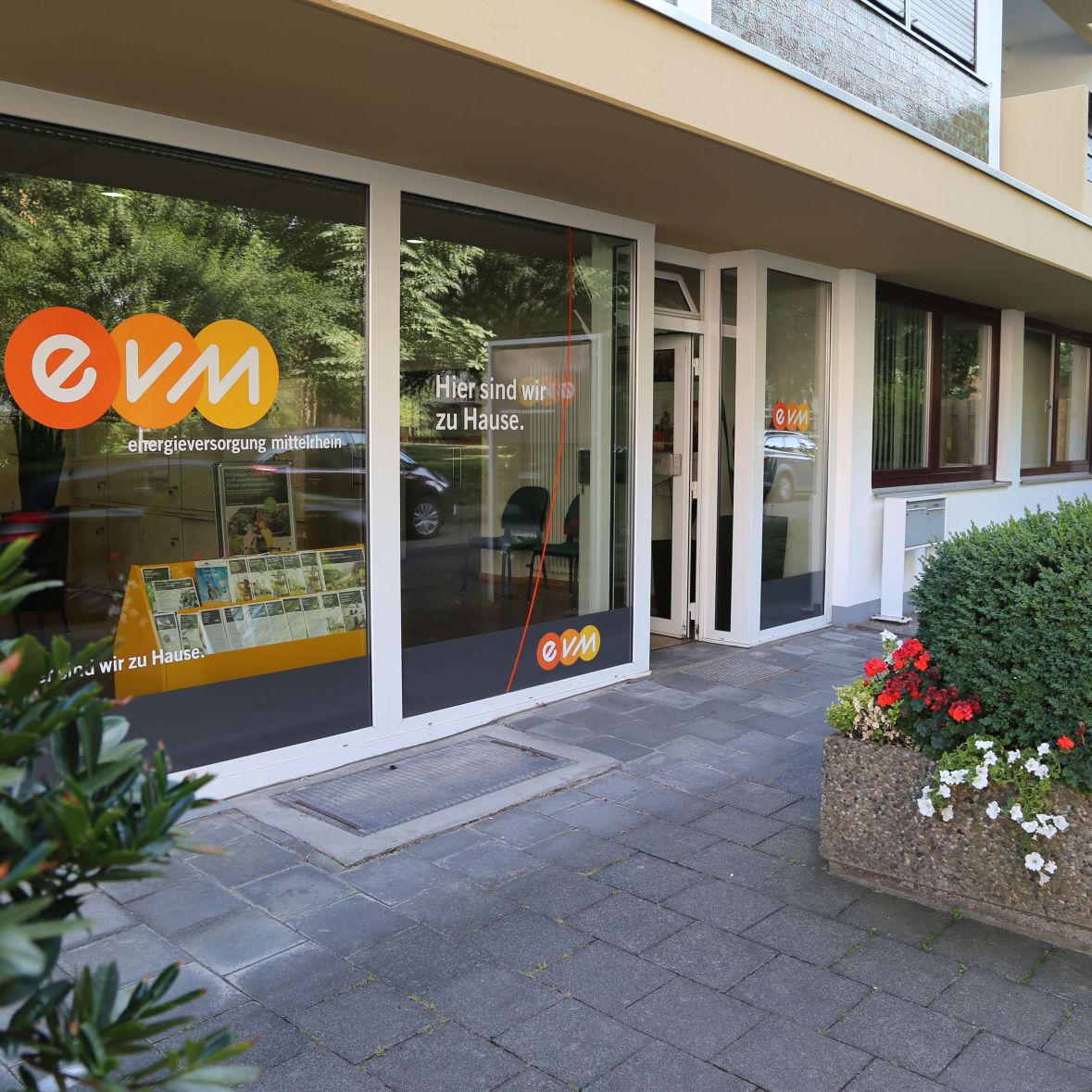 Ein Bürogebäude mit großer Glasfront zeigt das Logo "evm" und den Text "Hier sind wir zu Hause. energieversorgung mittelrhein". Im Vordergrund: Pflastersteine und ein Blumenbeet.