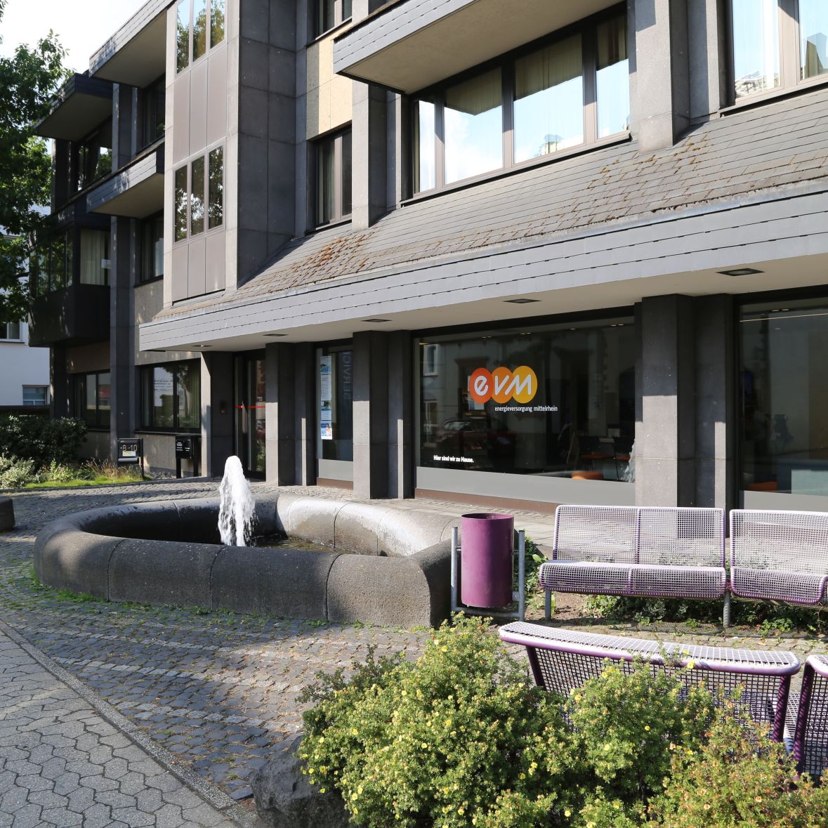 Brunnen sprudelt Wasser vor modernem Bürogebäude mit Glasfassade. Daneben stehen lila Mülleimer und Bänke. Im Hintergrund trägt ein Fenster das Logo "evm" in Orange und Weiß.