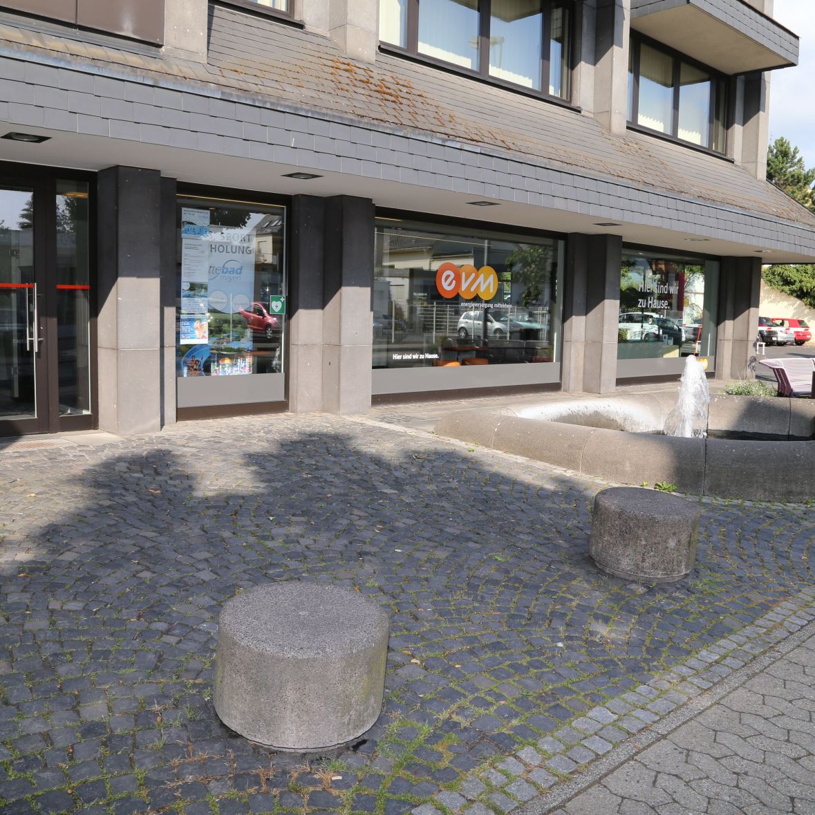 Ein Brunnen sprudelt Wasser auf gepflastertem Platz vor einem Gebäude mit großen Glasfenstern. Beschriftung auf der Scheibe: "evm", "Hier sind wir zu Hause". Im Hintergrund geparkte Autos und Bäume.