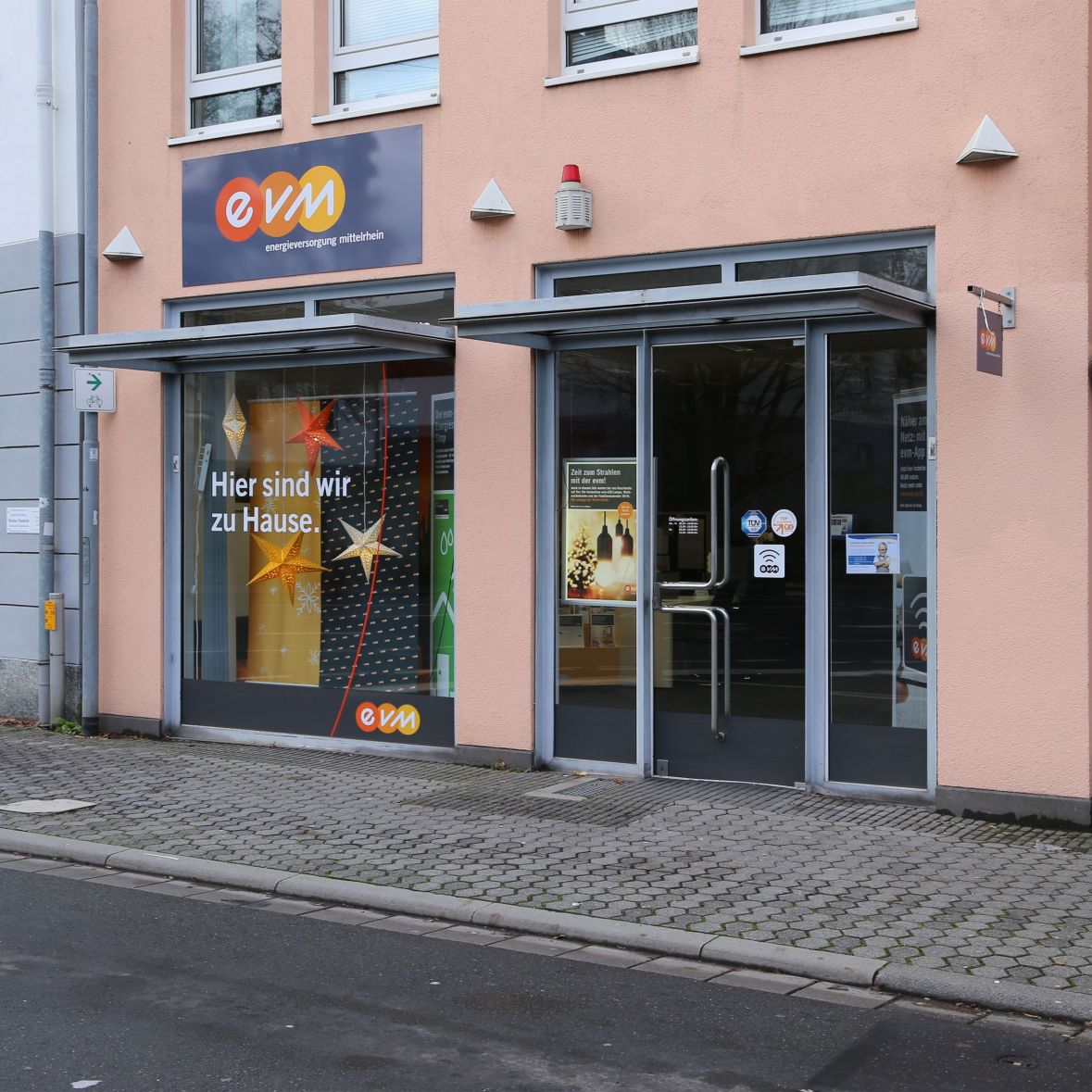 Ein Bürogebäude mit dem Schild "evm - energieversorgung mittelrhein", Fenster zeigen Dekorationen mit Text "Hier sind wir zu Hause", umgeben von einem Bürgersteig mit Pflastersteinen.