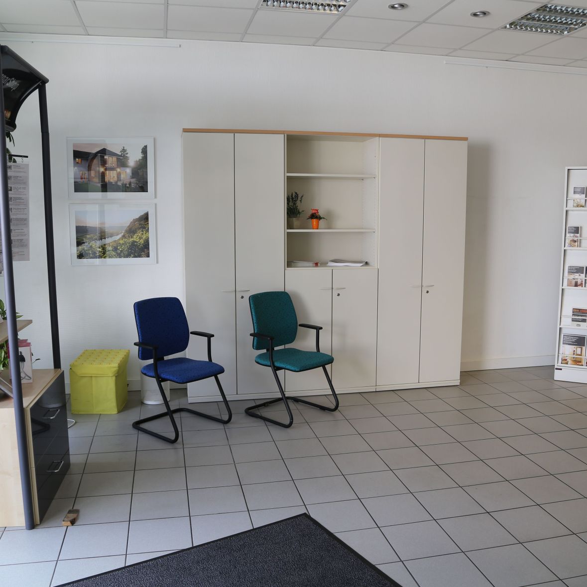 Ein Büro mit Bücherregal links und Broschürenhalter rechts. Zwei Stühle stehen vor einem weißen Schrank. An der Wand hängen Bilder. Pflanzen und ein gelber Würfel ergänzen die Einrichtung.