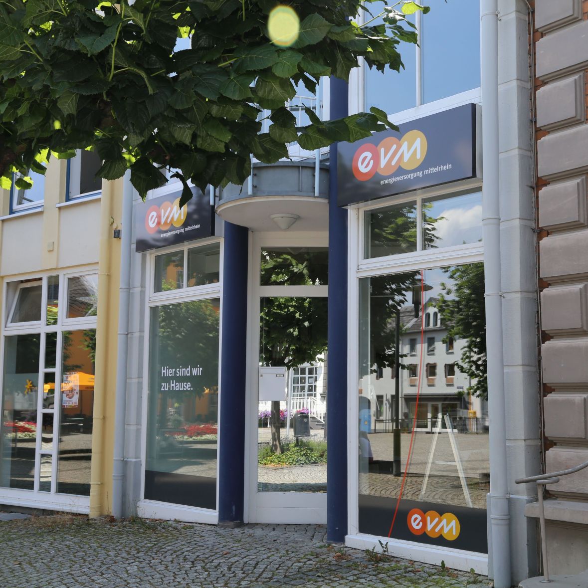 Ein Geschäftseingang mit großen Fenstern trägt das Logo "evm". Auf dem linken Fenster steht: "Hier sind wir zu Hause." Davor steht ein Baum. Im Hintergrund sind Gebäude erkennbar.