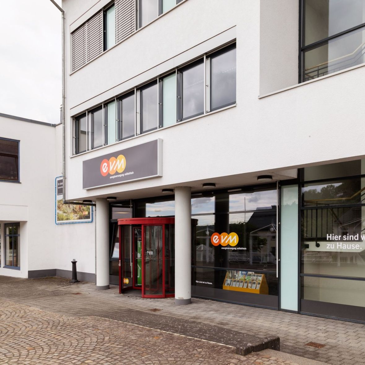 Ein Bürogebäude mit einer gewölbten Glasfront, der Eingang hat ein rotes Drehkreuz. Über dem Eingang ist ein Schild mit dem Logo "evm". Text: "Hier sind wir zu Hause."